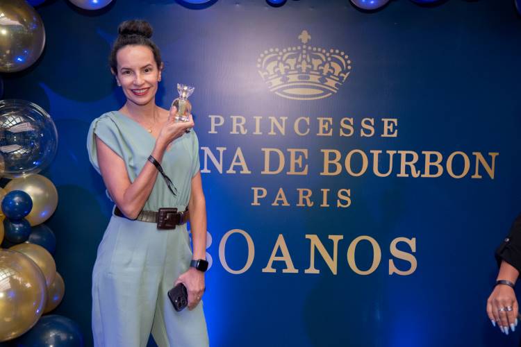 Mulher sorridente segura um frasco de perfume em um evento comemorativo. Atrás, um painel azul com a inscrição "Princesse Marina de Bourbon Paris - 30 Anos" e coroa dourada. Balões dourados e azuis decoram o ambiente elegante e festivo.
