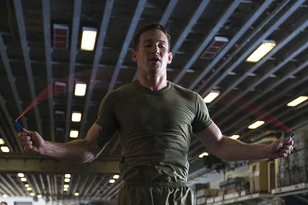 Homem de camiseta verde pulando corda em um hangar coberto, com os braços abertos no giro