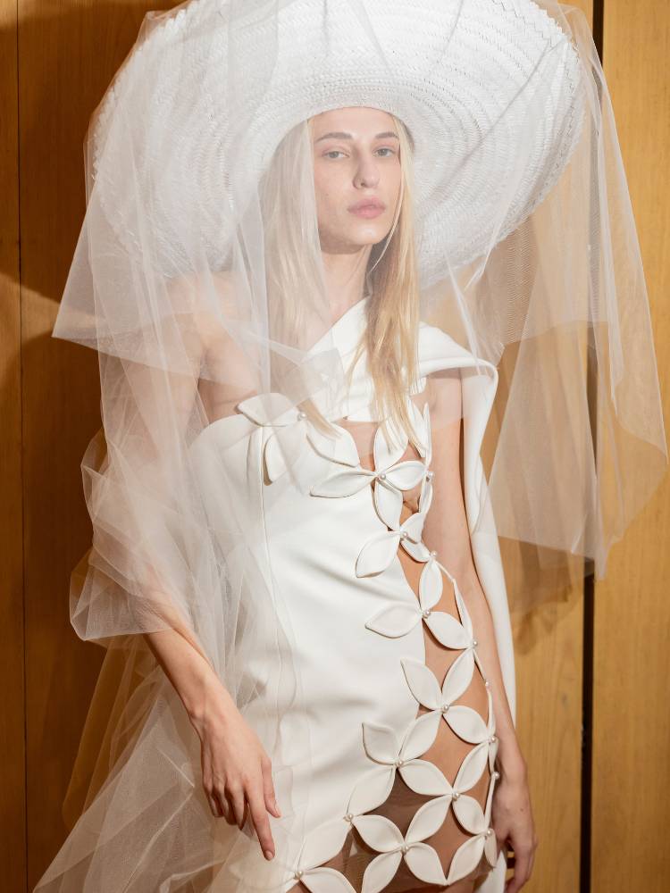 Mulher loira de pele clara usando chapéu grande branco, véu e vestido de noiva branco curto com recortes