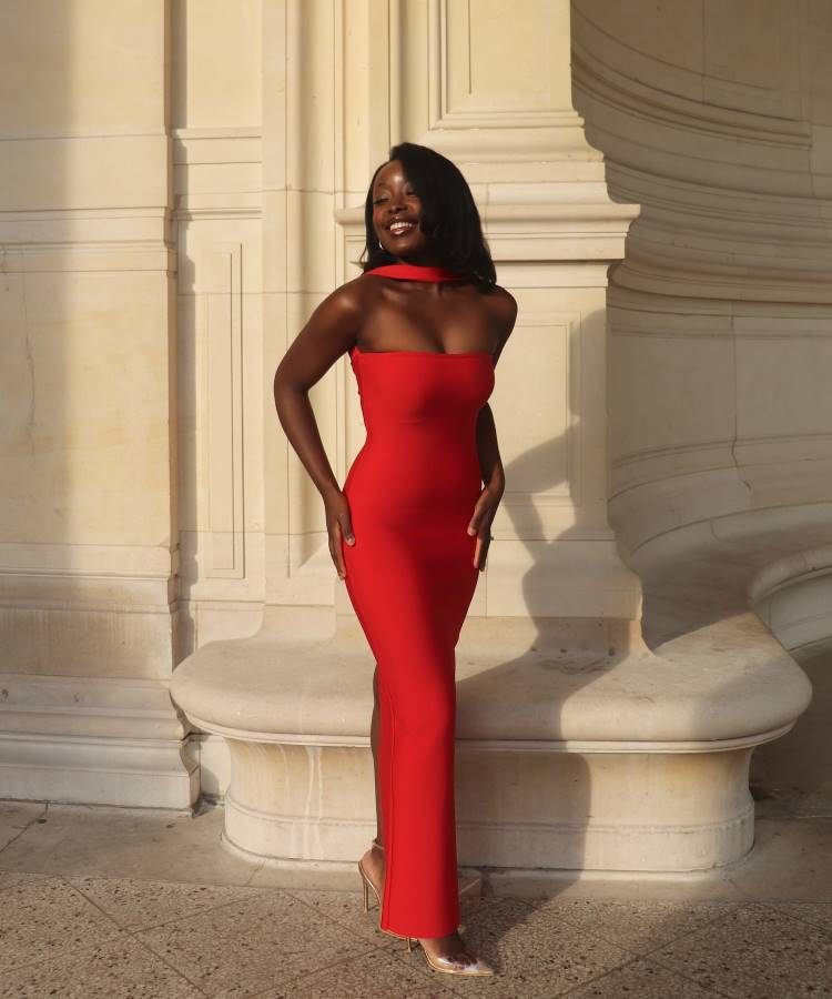 Uma mulher sorri, posando ao ar livre ao lado de um pilar de pedra. Ela usa um vestido vermelho justo, sem alças e com gola alta, sapatos de salto alto nude e brincos de argola dourados.







