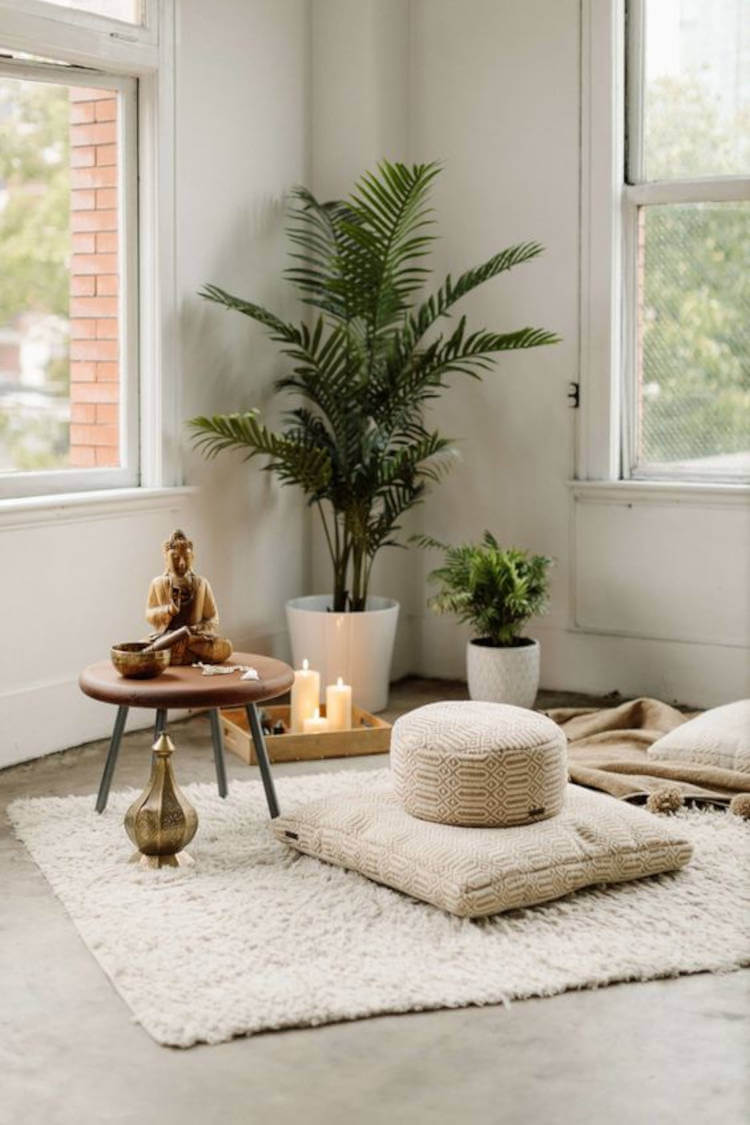 Espaço relaxante com tapete bege, almofadas, banquinho de madeira, estátua de Buda, vasos de planta, velas e janela