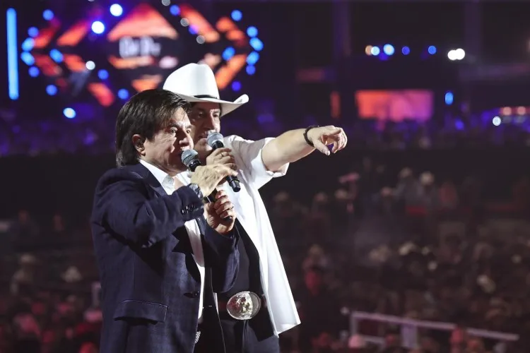 Dois homens em um palco iluminado, com luzes vibrantes ao fundo. O da esquerda, cabelo escuro, usa terno; à direita, chapéu cowboy e camisa branca. Ambos seguram microfones. "Nome Verdadeiro dos Famosos".
