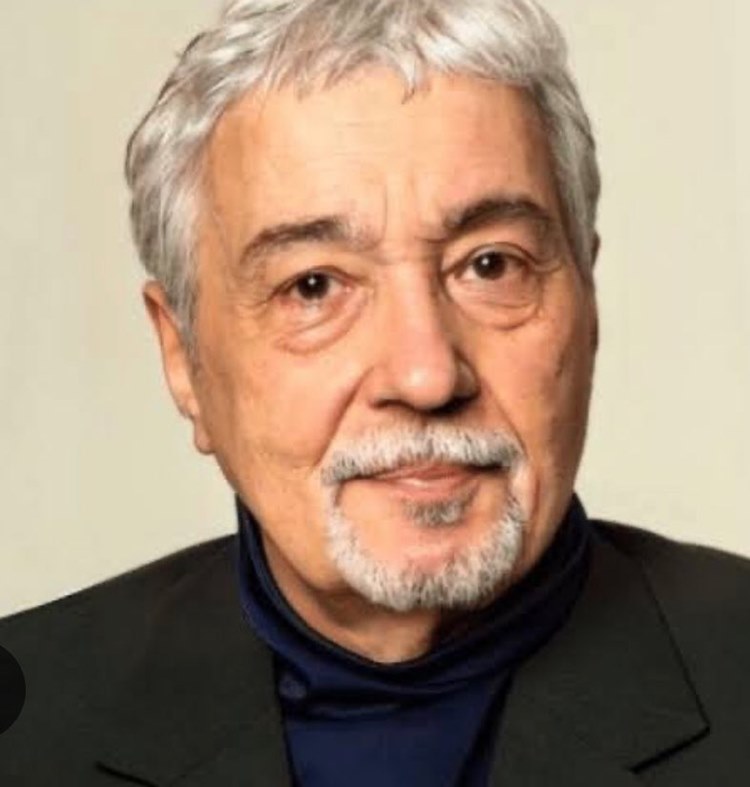 Um retrato de um homem de idade com cabelo grisalho e cavanhaque. Ele usa um blazer escuro sobre uma blusa de gola alta azul-marinho. Ele olha calmamente para a câmera contra um fundo claro e liso.