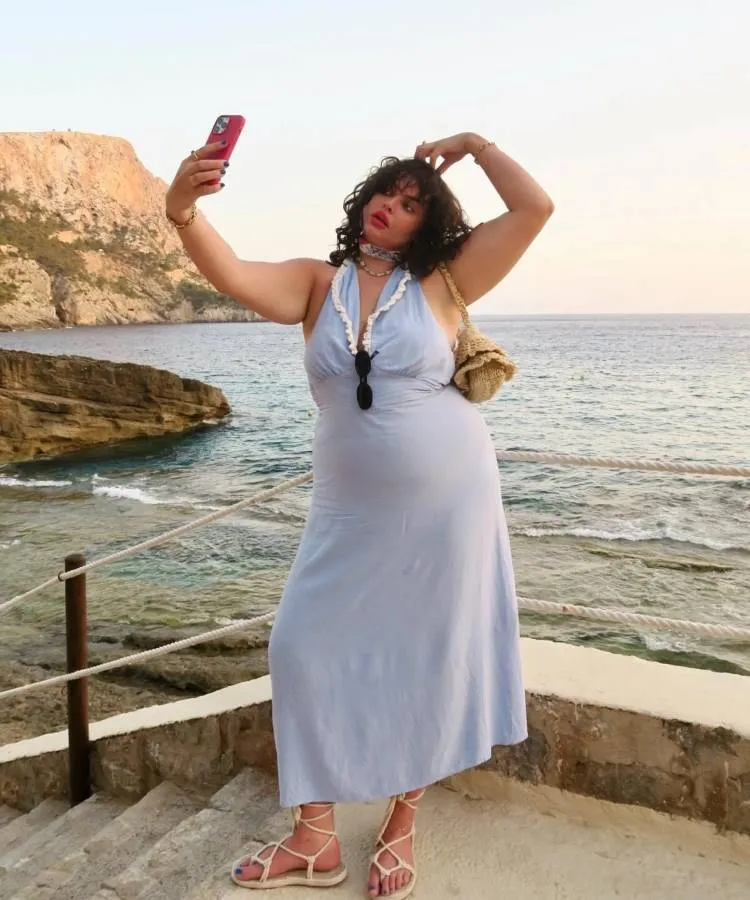 Uma mullher de vestido azul claro posa com um celular como se estivesse tirando uma selfie. Ela usa um vestido leve e sapato de amarração. Ao fundo está o mar.