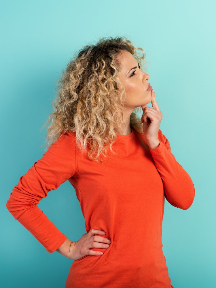 Mulher de pele clara usando blusa laranja, de lado, fazendo expressão de dúvida