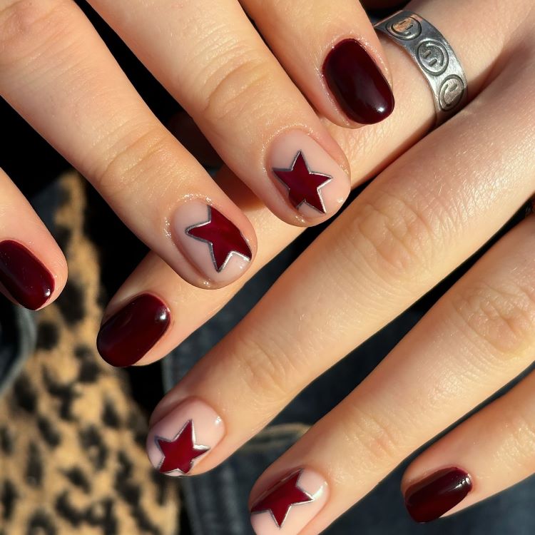 Mãos exibindo estilos de unhas curtas, decoradas com esmalte vinho brilhante e estrelas em destaque sobre base nude, contornadas por linhas prateadas. Fundo desfocado em tons quentes e animal print, valorizando o design elegante e criativo das unhas.