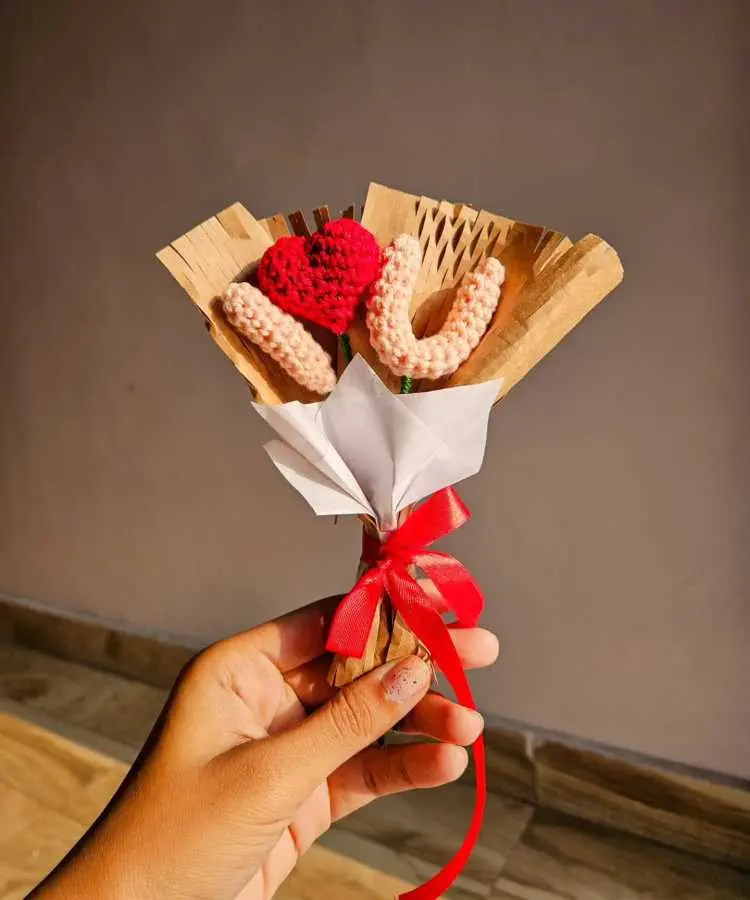 Um delicado buquê artesanal composto por flores de crochê em tons de rosa e um coração vermelho, envolto por folhas de papel kraft e laço vermelho vibrante. O buquê é segurado por uma mão contra um fundo neutro.