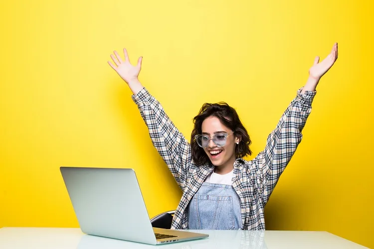 mulher de pele clara feliz, com os braços para cima, em frente ao notebook e com parede amarela atrás