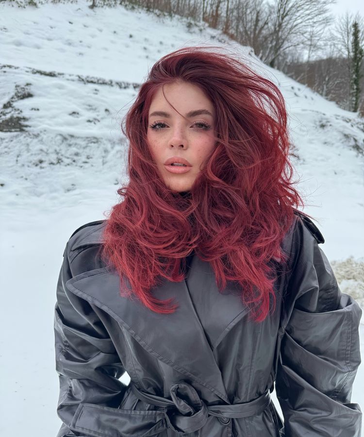 Mulher com cabelo ruivo volumoso, vestindo casaco preto de couro com cinto ajustado. Cenário de inverno com neve cobrindo o chão e árvores ao fundo, criando uma atmosfera fria e contrastante com seu visual marcante.