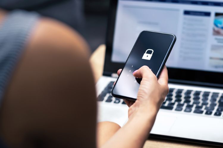 Mulher segurando telefone e digitando a sua senha de segurança, em frente a um notebook com tela embaçada