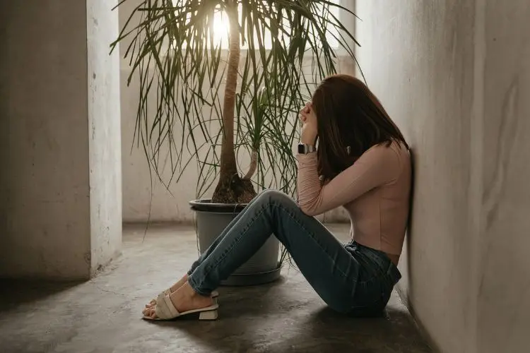Uma mulher senta-se no chão encostada a uma parede, com as mãos cobrindo o rosto em um momento de angústia. Ao lado, um vaso com uma planta e luz natural entrando pela janela criam um contraste emocional.