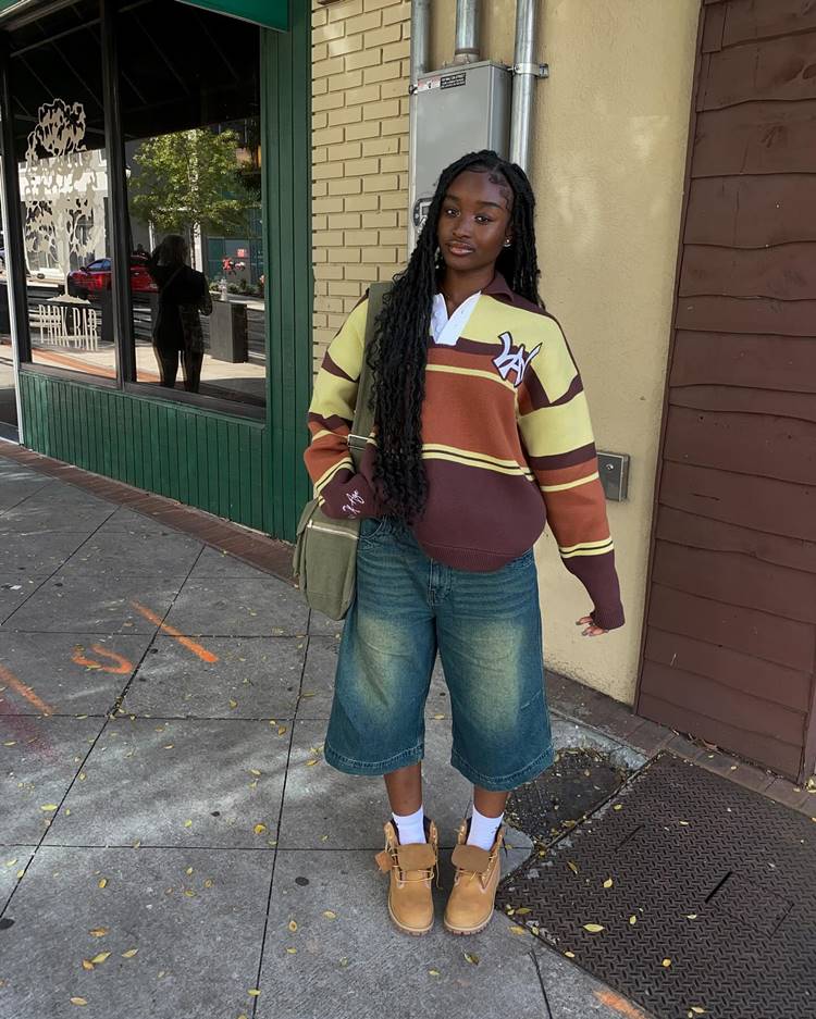 Garota negra usando jorts, sapato caramelo com meia branca cano alto de moletom listrado, bolsa verde, em local público.