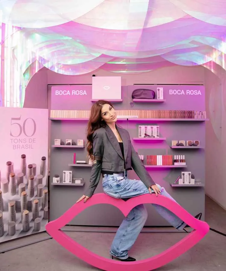 Bianca Andrade posando em estande da marca Boca Rosa, decorado em tons rosa, com produtos de maquiagem expostos. Ela usa blazer cinza e jeans, sentada em escultura rosa em forma de lábios. Iluminação colorida ao fundo.