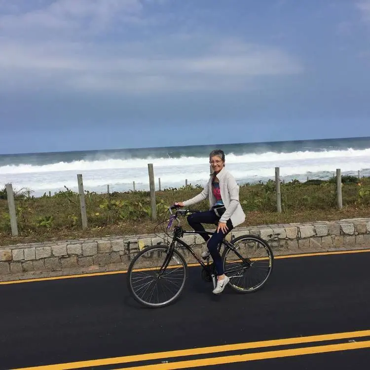 Mulher sorridente sentada em uma bicicleta preta sobre asfalto com faixas amarelas. Ao fundo, uma cerca, vegetação rasteira e o mar com ondas brancas sob céu parcialmente nublado. Cena transmite tranquilidade e lazer ao ar livre.
