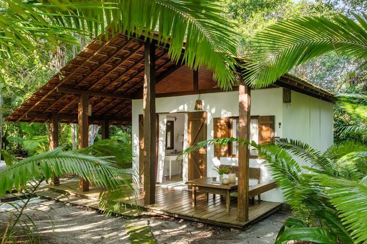 Imagem de fachada de casa simples, com alpendre, que mostra uma casinha branca com uma mesa do lado de fora e muitas plantas e árvores ao redor