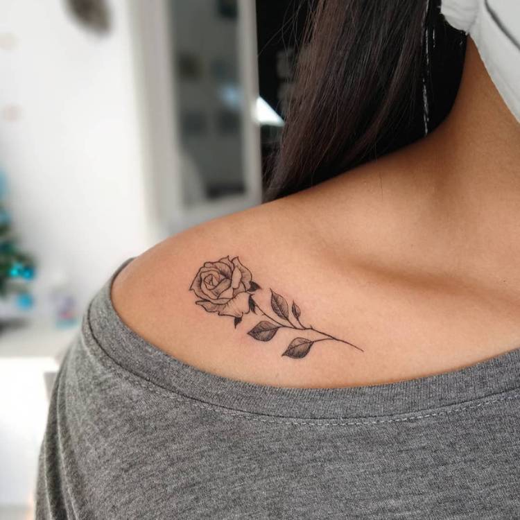 Mulher de pele morena e blusa cinza com rosa tatuada em preto estilo fine line no ombro