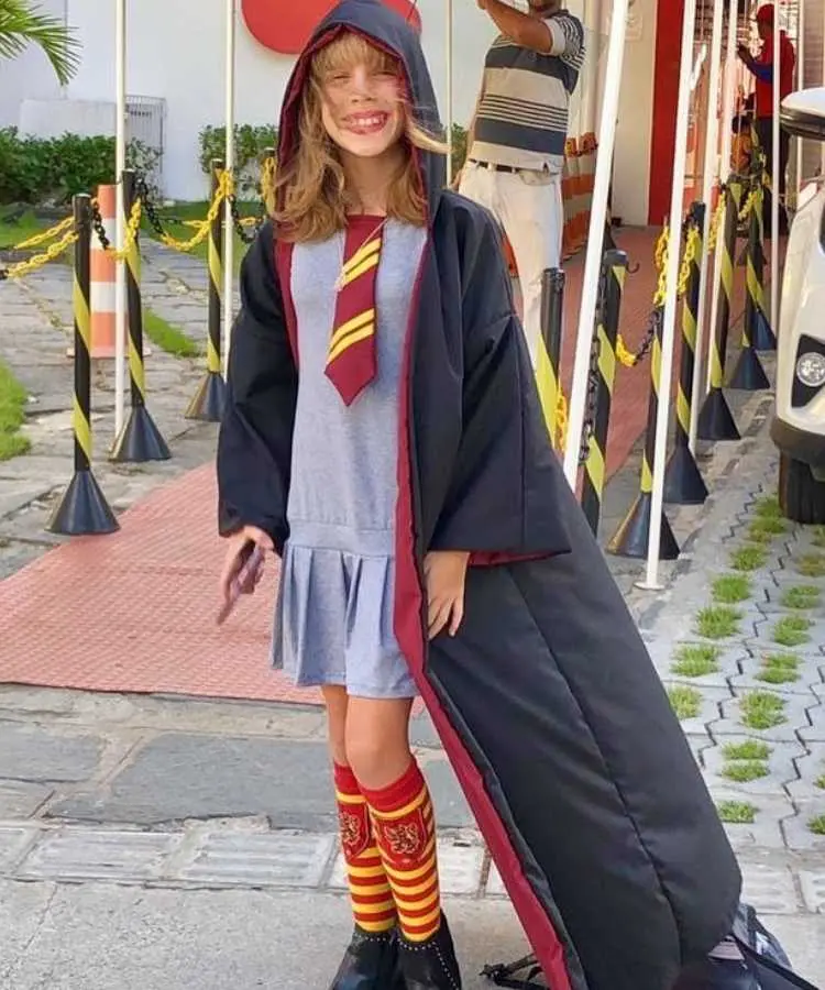 Garota com fantasia de bruxa infantil Harry Potter. Cores predominantes: cinza, preto, vermelho e amarelo. Usa: vestido com gravata, capa longa com touca, meias longas e sapato preto