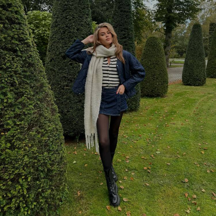 Uma mulher loira caminha por um jardim com arbustos verdes em forma de cone. Ela usa uma jaqueta acolchoada azul marinho, uma blusa listrada, minissaia escura, meias-calças pretas, botas e um cachecol claro, exalando um estilo aconchegante e chic de outono.