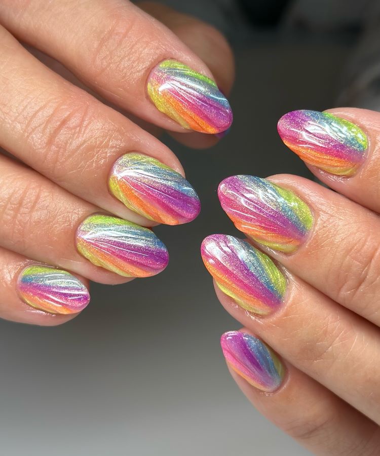 A imagem exibe unhas curtas e ovais decoradas com um design vibrante e furta-cor. As cores do arco-íris, como rosa, azul, amarelo e verde, são aplicadas em camadas onduladas e texturizadas, criando um efeito tridimensional brilhante.