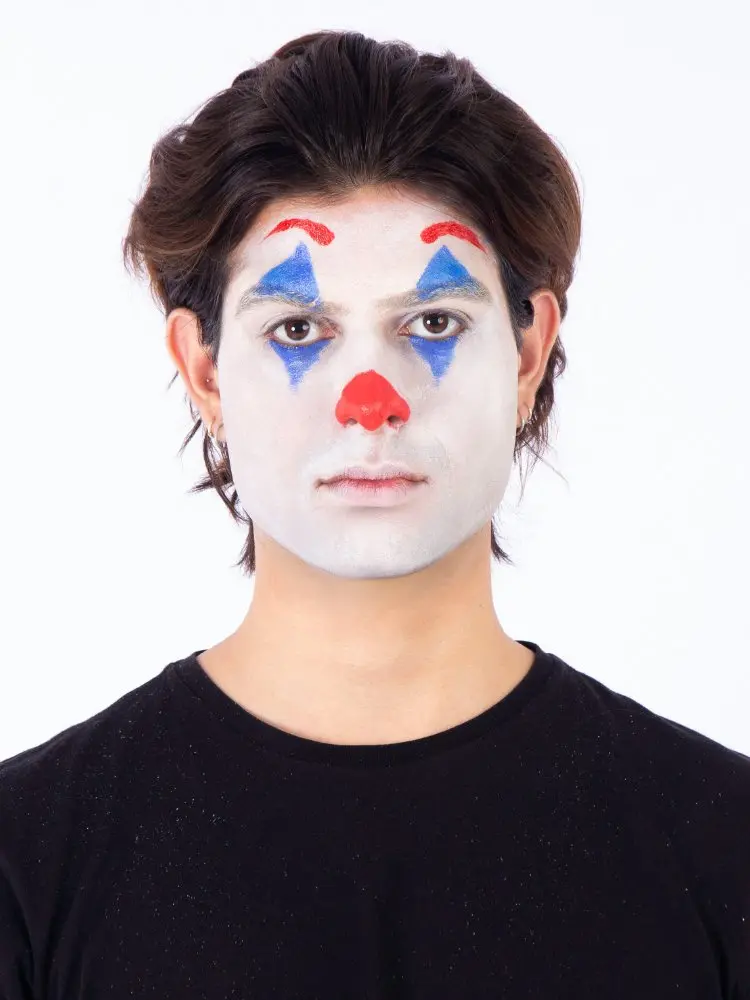 Homem de pele clara, cabelo médio castanho, usando blusa preta, pancake branco no rosto, clown azul nos olhos, clown vermelho na sobrancelha e no nariz 