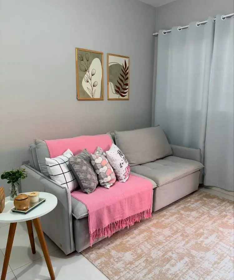 Uma sala aconchegante com sofá cinza, manta rosa e almofadas decorativas. Uma mesa lateral redonda branca exibe velas e um vaso pequeno. Duas molduras com motivos de folhas adornam a parede cinza, e cortinas claras completam o ambiente. Tapete em tons neutros no chão.







