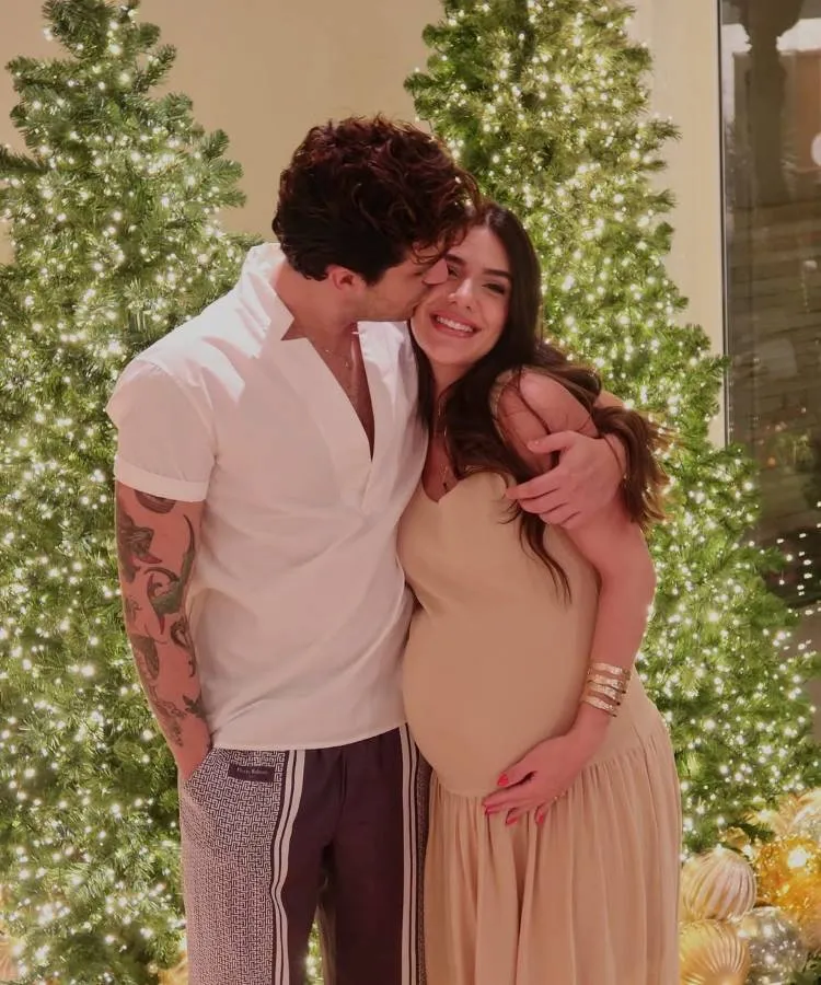 Esta imagem mostra Luan Santana com sua noiva, Jade Magalhães, que aparenta estar grávida, em um ambiente festivo com árvores de Natal iluminadas. Luan beija a testa de Izabela, que sorri e segura a barriga.

