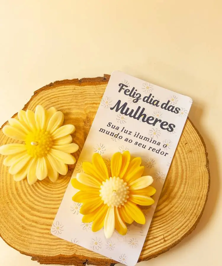 Cartão decorativo com a mensagem "Feliz dia das Mulheres - Sua luz ilumina o mundo ao seu redor" e flores artificiais amarelas. Exibido sobre uma base de madeira com fundo neutro, ideal como lembrança delicada para homenagear mulheres.