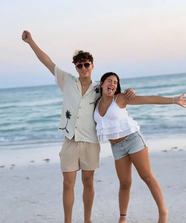 A imagem mostra dois jovens sorrindo em uma praia arenosa. O rapaz, à esquerda, veste camisa de palmeiras e shorts, com um braço erguido. A moça, à direita, usa top branco e shorts jeans, com a língua para fora e braços abertos, em frente ao oceano.







