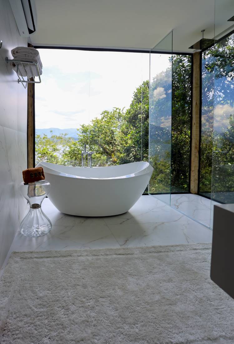 Banheira moderna e branca em um banheiro minimalista, com grandes janelas que oferecem uma vista deslumbrante da natureza e do mar. O piso e as paredes são de mármore claro.







