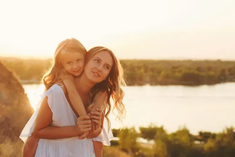 Mãe com vestido branco carrega sua filha loira sorridente nas costas em um cenário ao ar livre, com luz dourada do pôr do sol e um rio ao fundo. O momento captura amor, serenidade e uma conexão familiar especial.