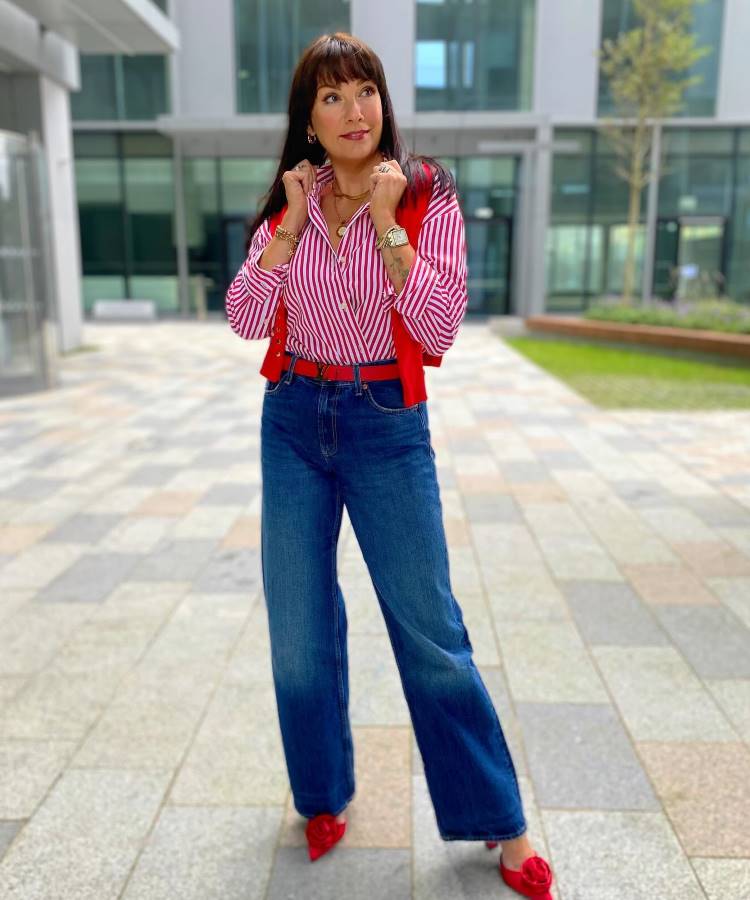 A imagem apresenta uma calça jeans wide leg, tendência para 2025, com lavagem azul clássica e cintura alta. O modelo destaca-se pelo caimento solto e elegante, complementado por uma camisa listrada e sapatos vermelhos, criando um visual moderno e vibrante.