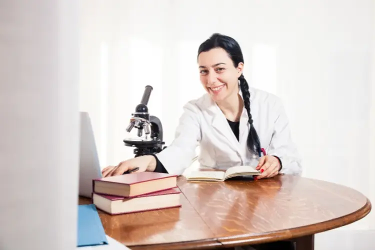 Mulher de pele clara à mesa com livros e microscópio usando jaleco