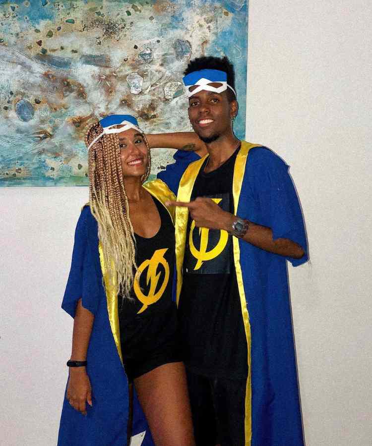 Fantasia de casal para o Halloween: Super Choque. Mulher e homem de pele negra usando roupa preta com símbolo de Super choque, máscara do personagem e capa azul e dourada