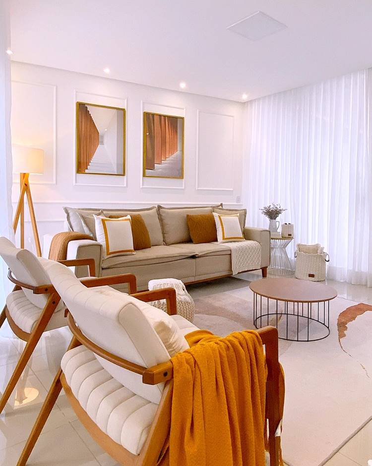 A imagem mostra uma sala de estar elegante e bem iluminada. Há um sofá grande e confortável no centro, adornado com almofadas brancas e douradas. Na frente do sofá, há uma mesa de centro redonda com tampo de vidro. Uma poltrona branca moderna com pernas de madeira está à esquerda, coberta por um cobertor amarelo mostarda. À direita do sofá, há uma pequena mesa lateral branca com uma planta. Três quadros retangulares estão pendurados na parede atrás do sofá; cada quadro contém uma peça abstrata de arte geométrica dourada sobre um fundo branco. À esquerda da imagem, há um tripé de madeira que sustenta uma lâmpada de assoalho moderna com uma sombra branca. Cortinas brancas longas cobrem a parede à direita, sugerindo a presença de janelas ou portas para o exterior. A sala é dominada por cores claras, com paredes brancas e um piso de cor clara.

