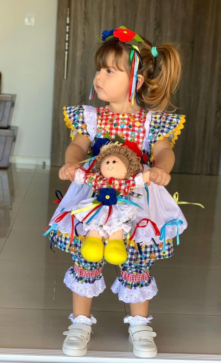 Menina de pele clara com vestido xadrez vermelho e azul segurando boneca com mesmo vestido