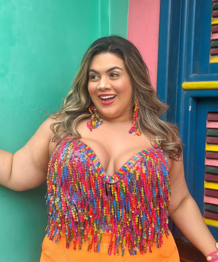 Mulher sorridente posa em frente a paredes coloridas azul, verde e rosa. Usa top com franjas de lantejoulas multicoloridas, brincos combinando e calça laranja vibrante. O ambiente alegre destaca um clima festivo e descontraído.
