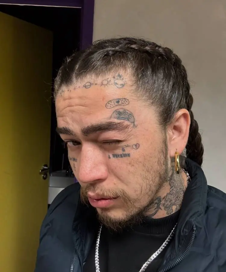 A imagem exibe o rosto de um homem com tranças nos cabelos e diversas tatuagens faciais, incluindo band-aids e símbolos. Ele possui barba e bigode finos, e usa um brinco de argola dourado e um colar prateado. Seu olhar está direcionado para a câmera, com um dos olhos levemente fechado.







