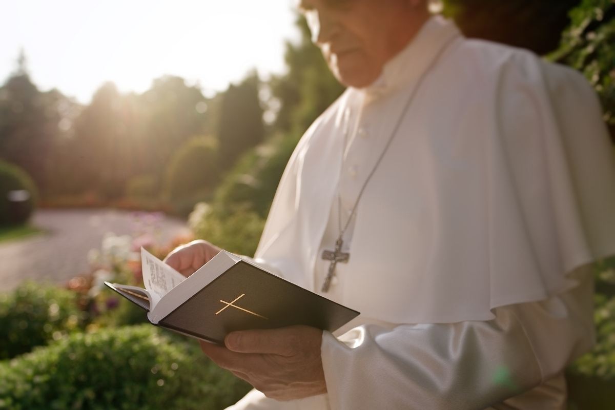 Um homem vestindo uma túnica branca e carregando um crucifixo lê um livro religioso em um jardim iluminado pelo sol, transmitindo serenidade e espiritualidade em meio à natureza e ao ambiente tranquilo ao seu redor.