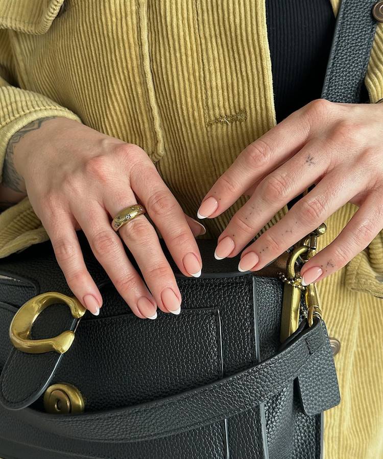 mulher usando casaco bege com bolsa preta de couro com detalhes dourados, ela está com as suas duas mãos segurando a bolsa que está em seu ombro, suas unhas elegantes possuem a decoração francesinha, ela usa um anel em uma mão e na outra tem tatuagens de pontos
