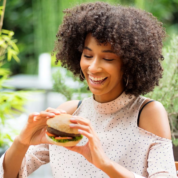 Mulher negra segurando um hámburguer em fundo desfocado com plantas