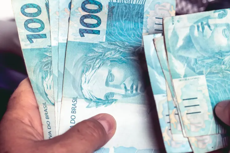 A imagem mostra mãos segurando várias notas de cem reais, exibidas em leque. As notas azuis destacam a efígie da República e detalhes em verde, simbolizando a moeda brasileira.


