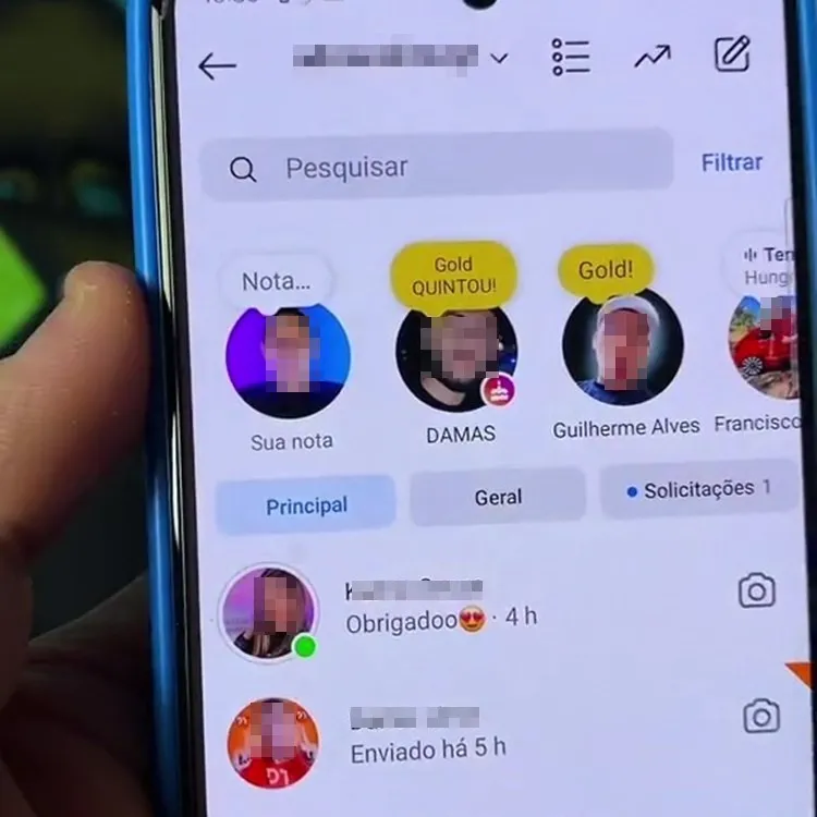 A imagem mostra a tela de um smartphone exibindo a interface de mensagens diretas do Instagram. No topo, há ícones de stories de diferentes usuários, alguns com etiquetas douradas que dizem "Gold QUINTOU!" e "Gold!". Abaixo, há uma barra de pesquisa e filtros, seguida por duas abas: "Principal" e "Geral". Duas mensagens recentes são visíveis, uma com a resposta "Obrigadoo?" enviada há 4 horas e outra "Enviado há 5 h". Uma mão segura o telefone, que está em uma capa azul.


