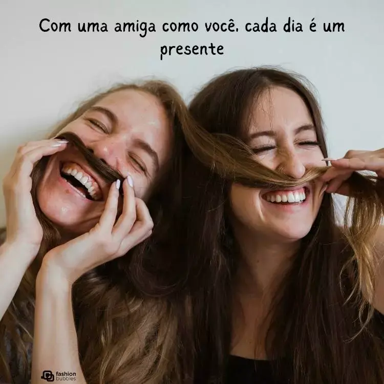 Duas mulheres de pele clara e cabelos castanhos sorrindo e usando mechas do cabelo como bigode. Acima dela, há a frase "Com um amigo como você, cada dia é um presente"