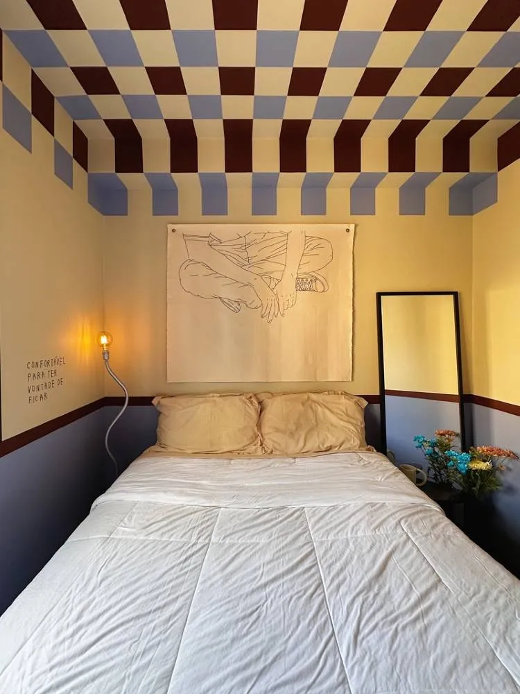 Esta é uma imagem de um quarto com cama de casal e roupa de cama clara. O teto e as paredes superiores têm um padrão geométrico em azul claro e bordô. Um espelho alto e uma luminária de piso com uma lâmpada exposta adicionam detalhes ao ambiente.

