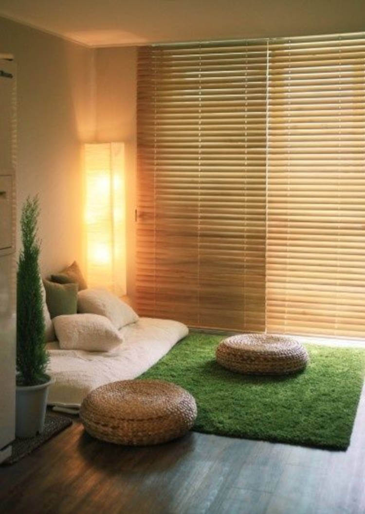 Espaço zen com tapete que imita grama, puffes beges, colchão com manta bege e almofadas felpudas, além de vaso com planta e persiana fechada