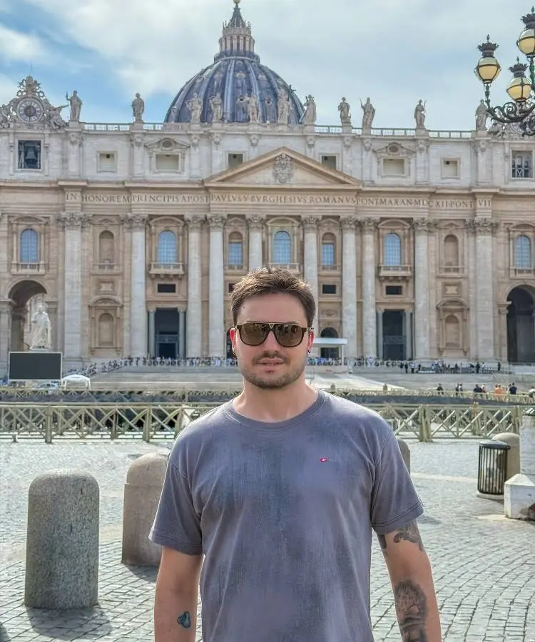 A imagem mostra um homem de óculos escuros e camiseta cinza, com tatuagens nos braços, em frente à majestosa Basílica de São Pedro no Vaticano. O céu está parcialmente nublado, e elementos arquitetônicos da basílica são visíveis ao fundo.







