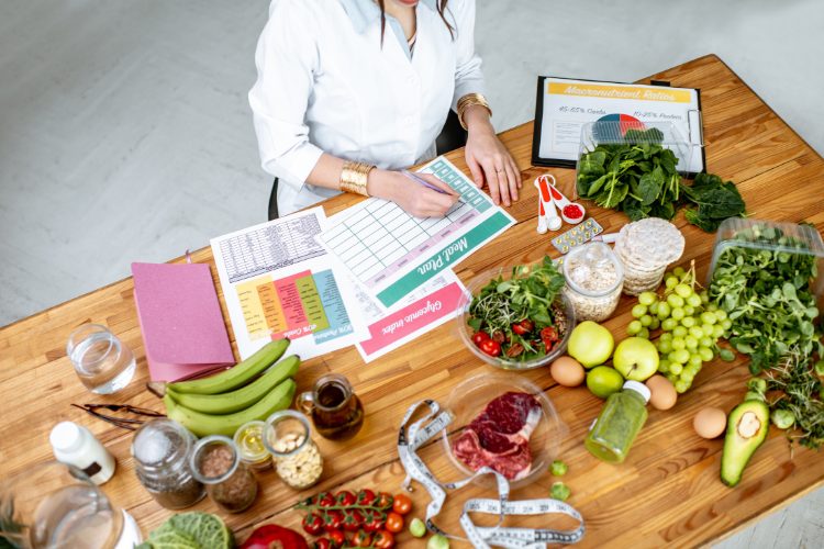 Foto tirada de cima de pessoa de pele clara usando jaleco, sentada à mesa montando plano alimentar com várias frutas e legumes ao redor