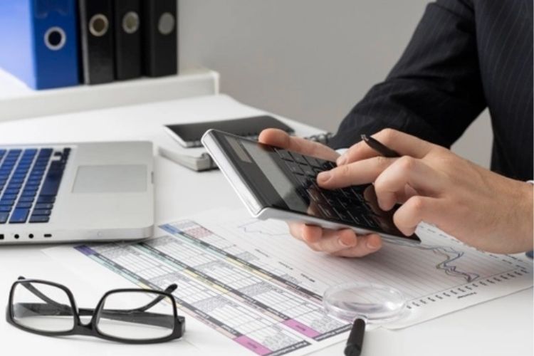 Foto de um homem fazendo conta na calculadora. Tem papéis, um óculos e um notebook em cima da mesa. crédito para compra de carro