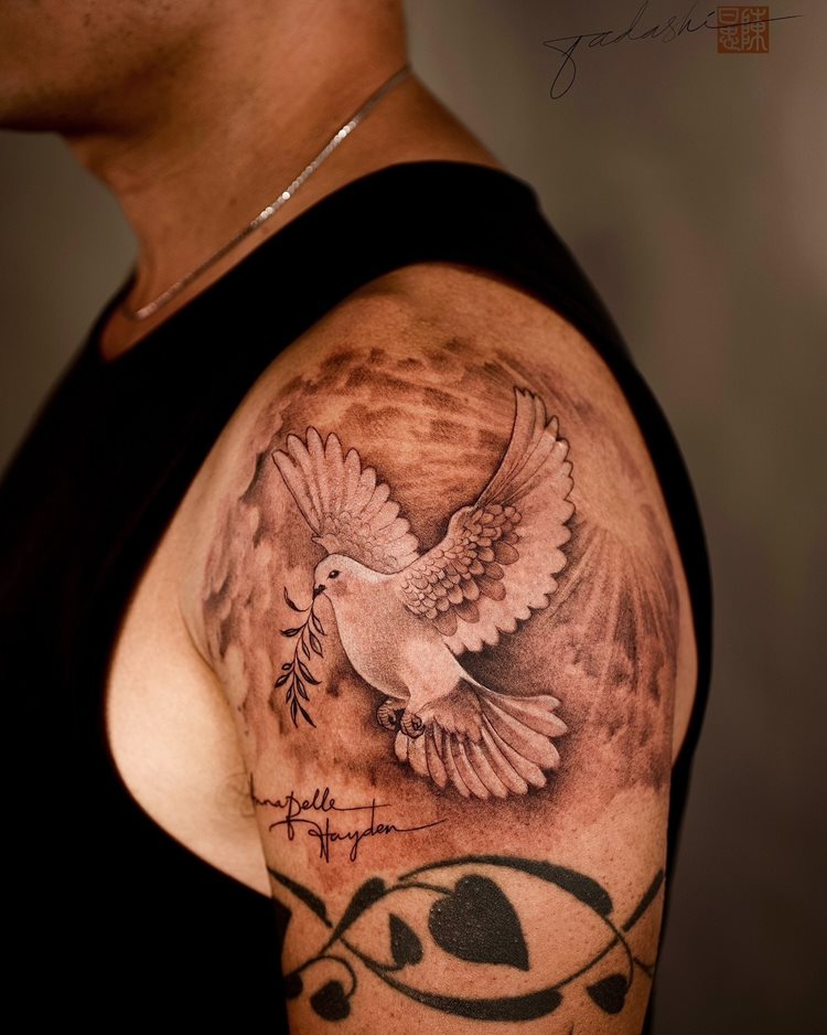 A imagem apresenta uma tatuagem religiosa no braço de um homem, destacando uma pomba em voo, simbolizando paz e espiritualidade. A pomba segura um ramo de oliveira, representando esperança. O fundo é sombreado, criando profundidade. Abaixo da pomba, há nomes escritos em estilo cursivo, adicionando um toque pessoal à arte.


