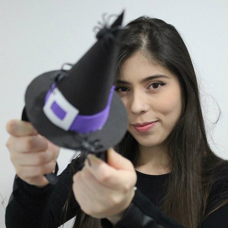 Mulher de pele clara usando blusa preta e segurando tiara com chapéu de bruxa